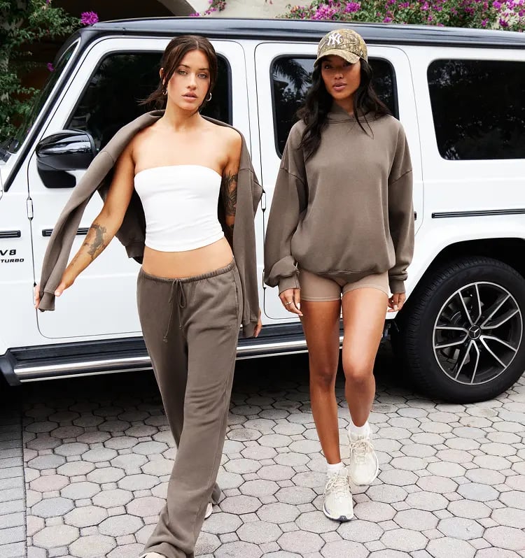 Two models walk out of the car. One is wearing a white tank top, taupe sweats, and a matching sweatshirt around her shoulders, the other is in a taupe hoodie and matching shorts. 