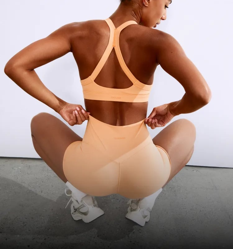 Model poses in a peach sports bra and matching booty shorts. 