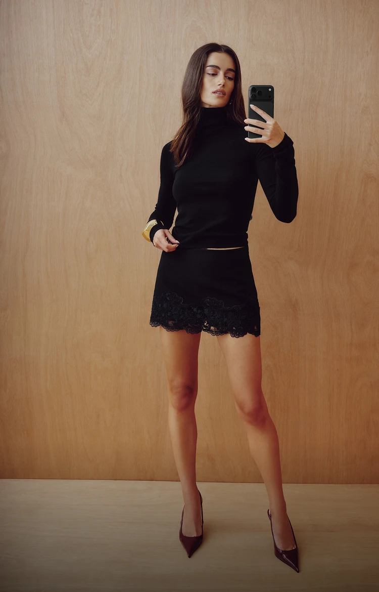 Model is wearing a black turtleneck and a black lace trim mini skirt.